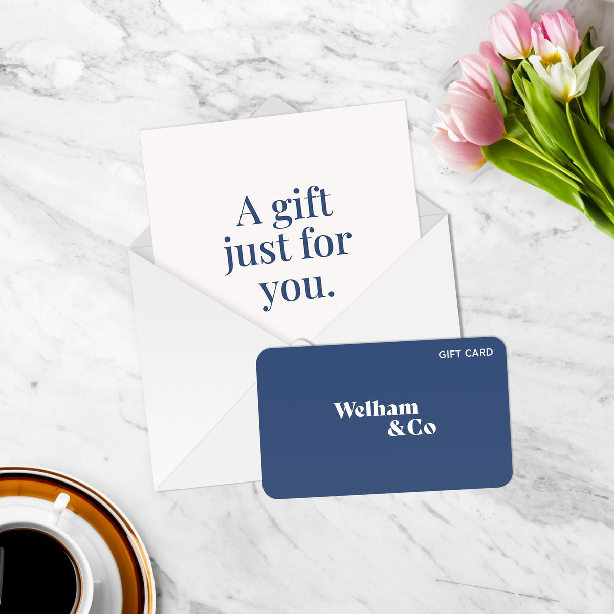 Navy blue Welham & Co gift card in front of an envelope and a card reading 'A gift just for you.', beside pink tulips