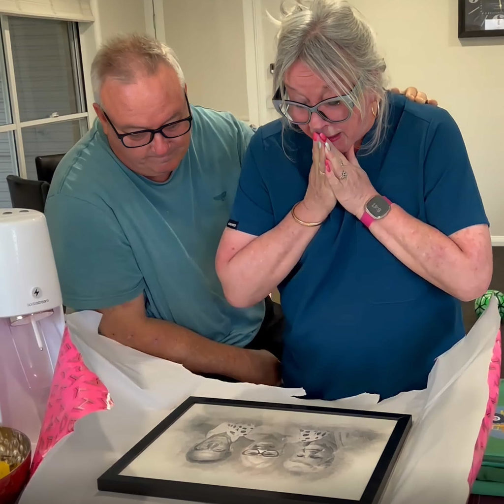 An older couple unwrap a black-framed sketch-style portrait; she presses her hands to her face