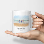 White mug held in a hand, printed with a watercolor-style portrait of a family walking into the sea at a beach