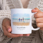 White mug held in two hands, printed with a watercolor-style portrait of a family at the shoreline