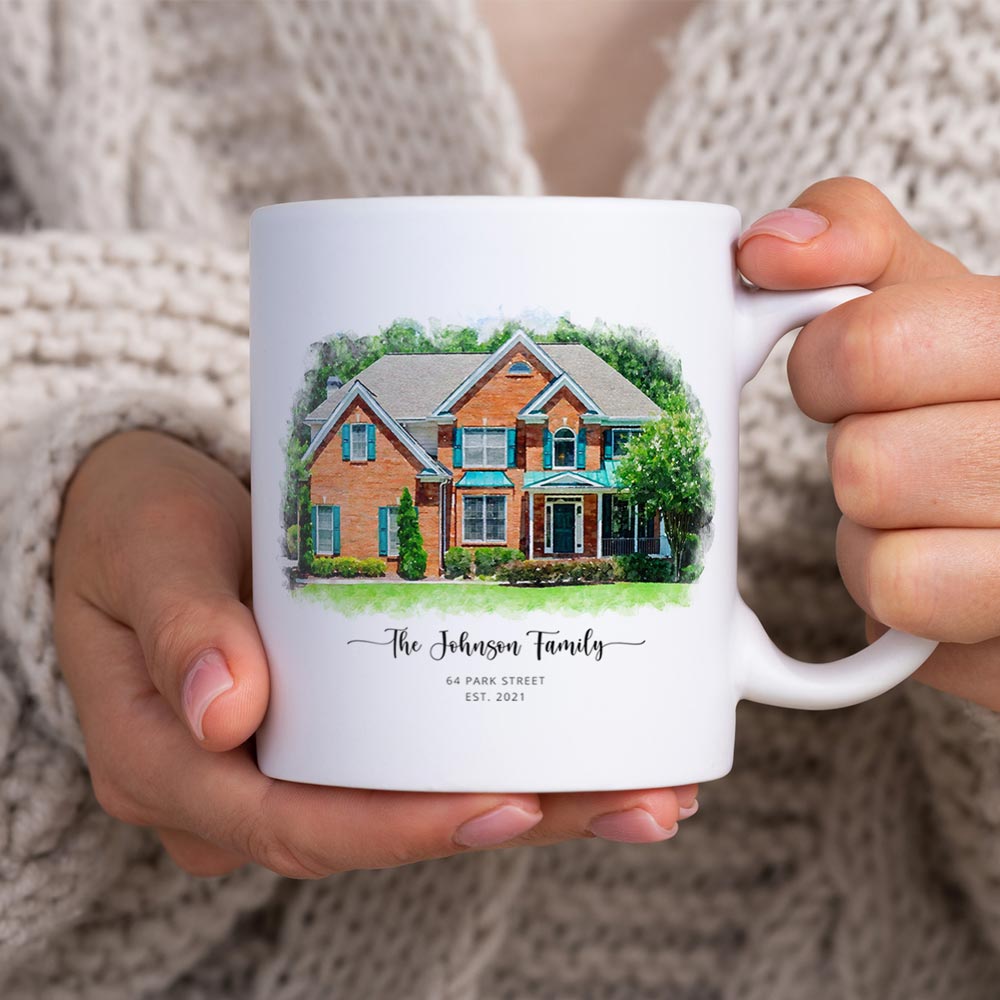 White mug held in two hands in a knit sweater, printed with a watercolor-style portrait of a red brick house